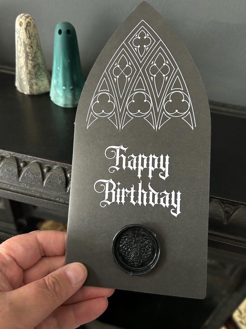 May include: A black birthday card with a gothic arch design and the words "Happy Birthday" in white script. The card has a black wax seal with a leaf design. Two ghost-shaped figurines are in the background.