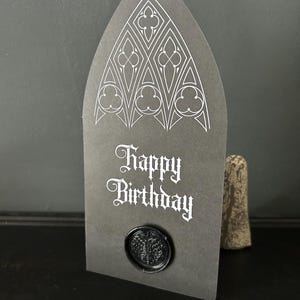 May include: A dark gray birthday card with a gothic arch design and the words "Happy Birthday" in white script. The card features a black wax seal. A decorative stone is in the background.