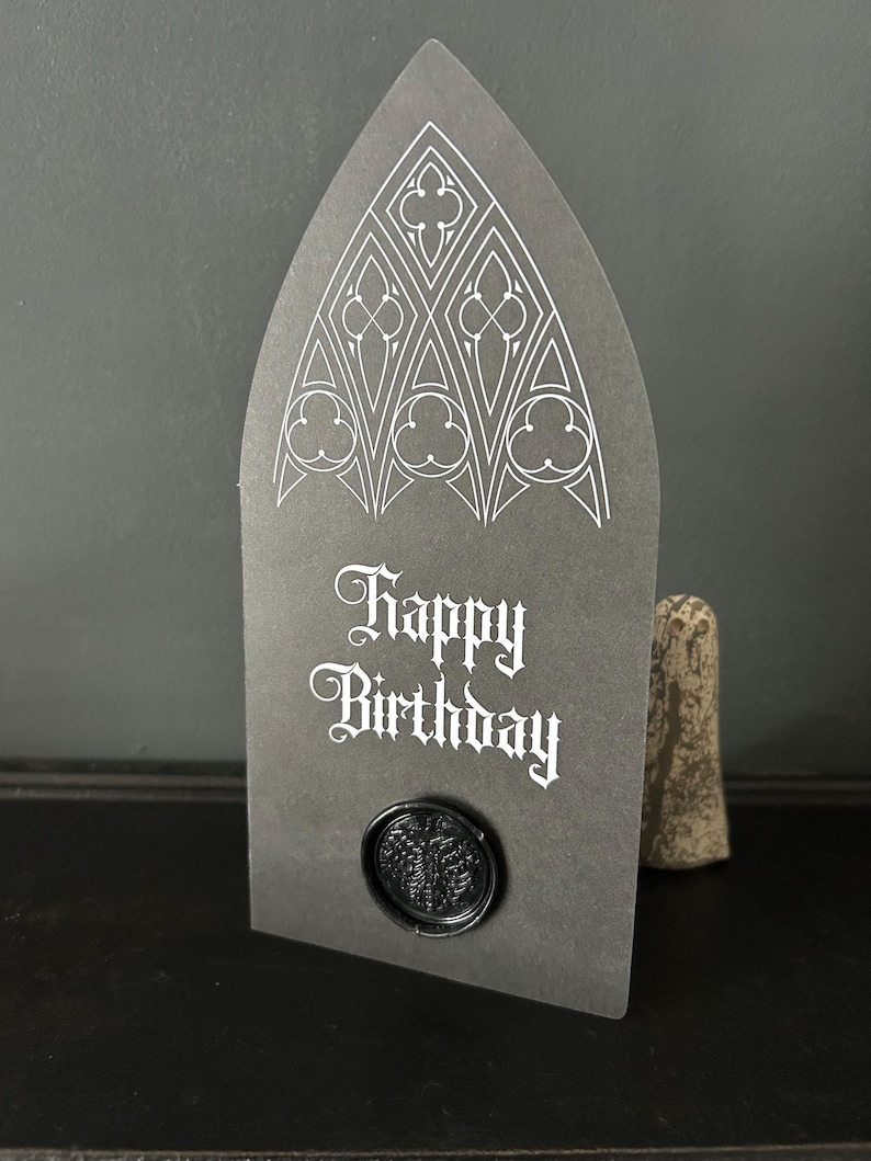 May include: A dark gray birthday card with a gothic arch design and the words "Happy Birthday" in white script. The card features a black wax seal. A decorative stone is in the background.