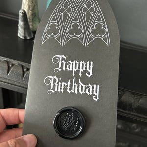 May include: A black birthday card with a gothic design and the words "Happy Birthday" in white script. The card is sealed with a black wax seal featuring a skeleton hand design.