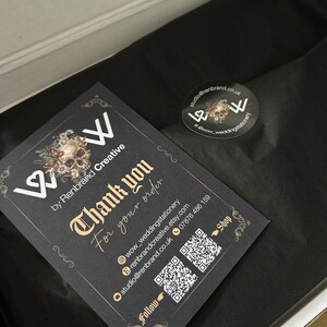 May include: A black card with gold text that reads "Thank you for your order" is placed inside a box with black tissue paper. A sticker with a skull design and the text "wow_weddingstationery" is also visible.