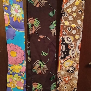 May include: Three long, narrow fabric strips hanging vertically. The left strip is bright blue with colorful floral patterns. The middle strip is dark brown with abstract green and orange starburst designs. The right strip features a brown, orange, and white paisley pattern.