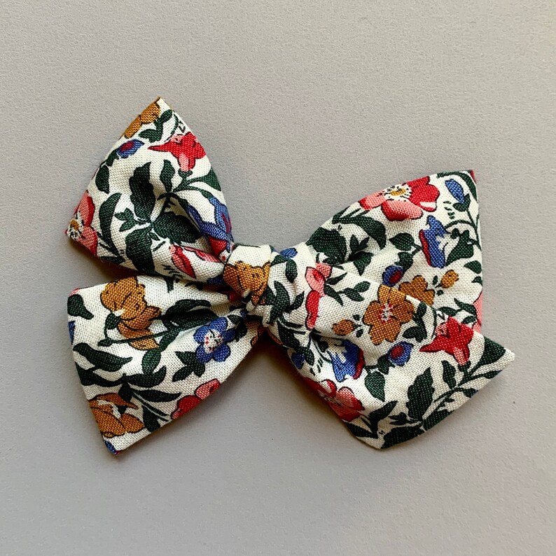 Puede incluir: Un lazo de tela blanco con un patr&oacute;n floral en tonos rojos, naranjas, azules y verdes. El lazo est&aacute; atado en una forma cl&aacute;sica con un nudo central.