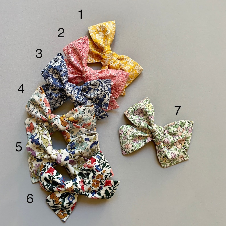 Puede incluir: Siete lazos para el pelo con estampado floral en varios colores y patrones. Los lazos est&aacute;n dispuestos en semic&iacute;rculo sobre un fondo gris.