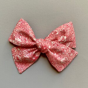 Puede incluir: Un lazo para el pelo de color rosa con estampado floral y flores blancas. El lazo est&aacute; anudado en el centro.