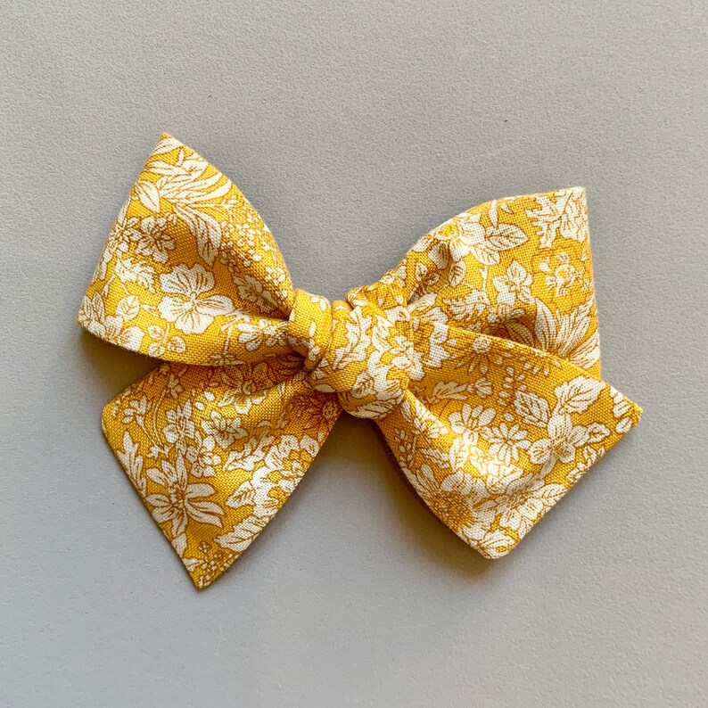Puede incluir: Un lazo de tela floral amarillo con flores y hojas blancas. El lazo est&aacute; atado en un nudo en el centro.
