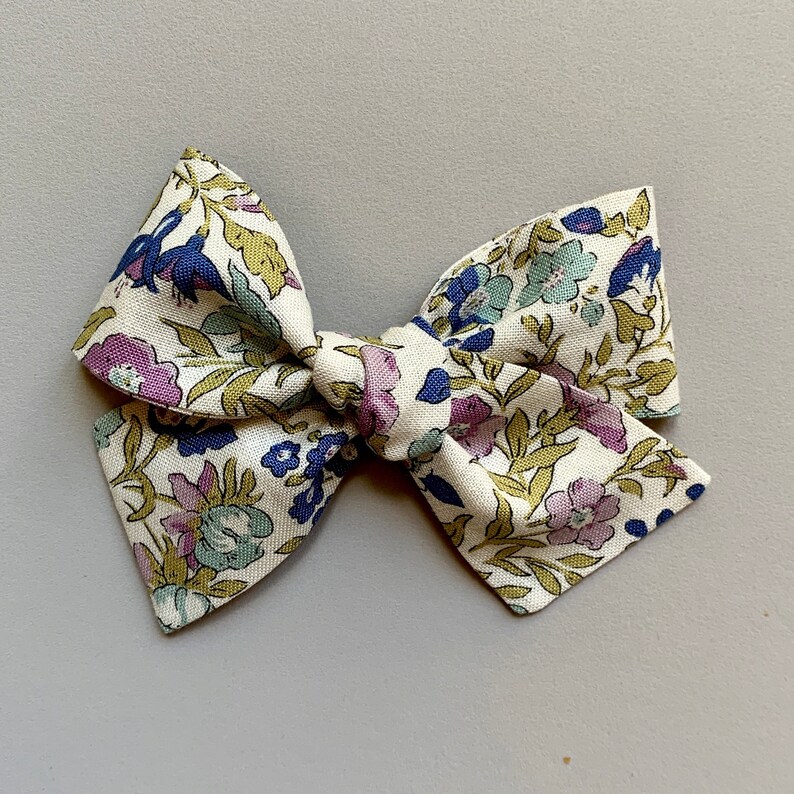 Puede incluir: Un lazo de tela blanco con un patr&oacute;n floral en tonos azules, verdes y morados. El lazo est&aacute; atado en un nudo y tiene una forma puntiaguda.