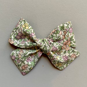 Puede incluir: Un gran lazo de color crema con un estampado floral en tonos rosa, amarillo y verde. El lazo est&aacute; hecho de tela y tiene un centro anudado.