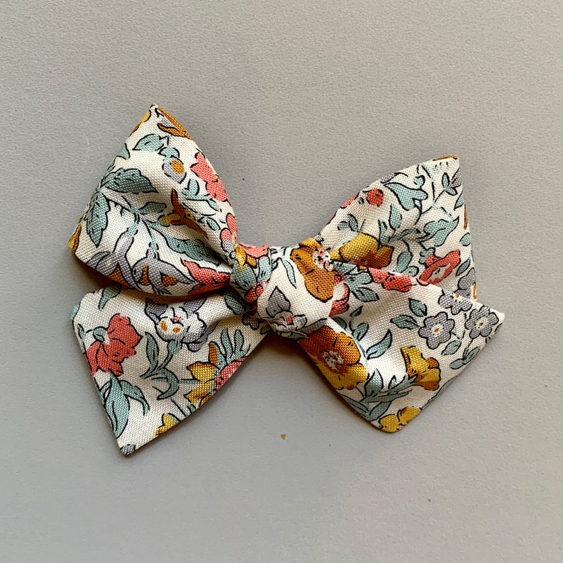 Puede incluir: Un lazo de tela blanco con un patr&oacute;n floral en tonos de rosa, amarillo y azul. El lazo est&aacute; atado en un nudo y tiene un borde ligeramente deshilachado.