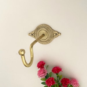 May include: A gold-toned metal hook with an ornate design. The hook is attached to a white wall.