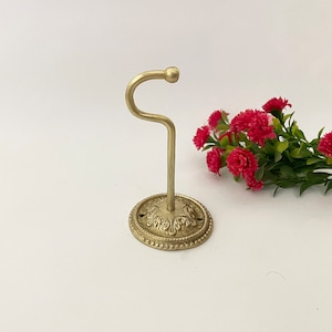 Pode incluir: Um gancho de metal dourado com uma base adornada. O gancho está preso a uma base redonda com um design decorativo.