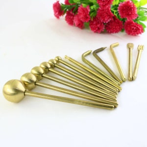 May include: A set of ten brass tools with rounded and pointed tips. The tools are arranged in a fan shape on a white surface.