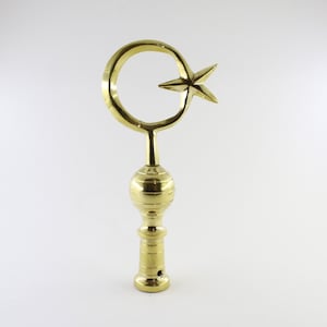 Puede incluir: Un remate de metal dorado con un diseño de media luna y estrella. El remate tiene una base redonda y un eje cilíndrico.
