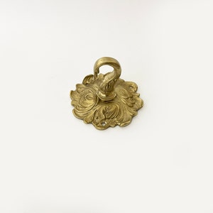 May include: A gold-toned metal ceiling hook with an ornate, floral design. The hook is designed to be mounted to a ceiling and has a decorative, scrolled hook.