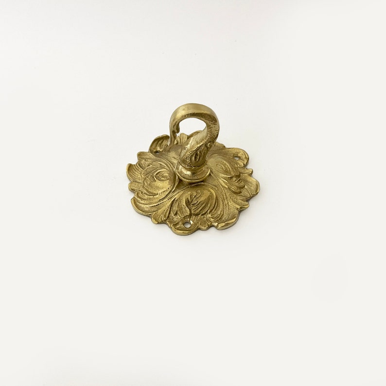 May include: A gold-toned metal ceiling hook with an ornate, floral design. The hook is designed to be mounted to a ceiling and has a decorative, scrolled hook.