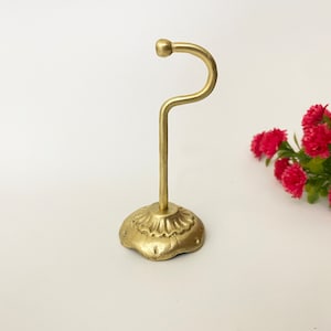 Puede incluir: Un soporte de gancho de metal dorado con una base decorativa. El gancho es curvo y tiene una parte superior redondeada. La base tiene forma de flor con un borde festoneado.