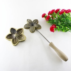 Op de afbeelding: Twee stervormige mallen van messing om bloemvormige lekkernijen te maken. Een mal is bevestigd aan een houten handvat en een metalen staaf. De mallen hebben een gedetailleerd bloemmotief.