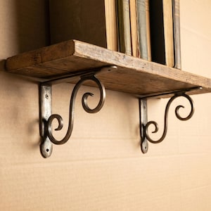 May include: A wooden shelf with decorative metal brackets. The shelf is made of reclaimed wood and supported by two ornate, scroll-design brackets. Several antique books are placed on the shelf, creating a vintage aesthetic.