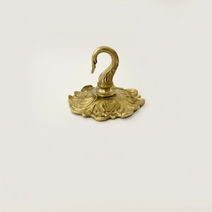 May include: A gold-toned metal hook with a decorative swan design. The hook is attached to a circular base with a floral pattern.