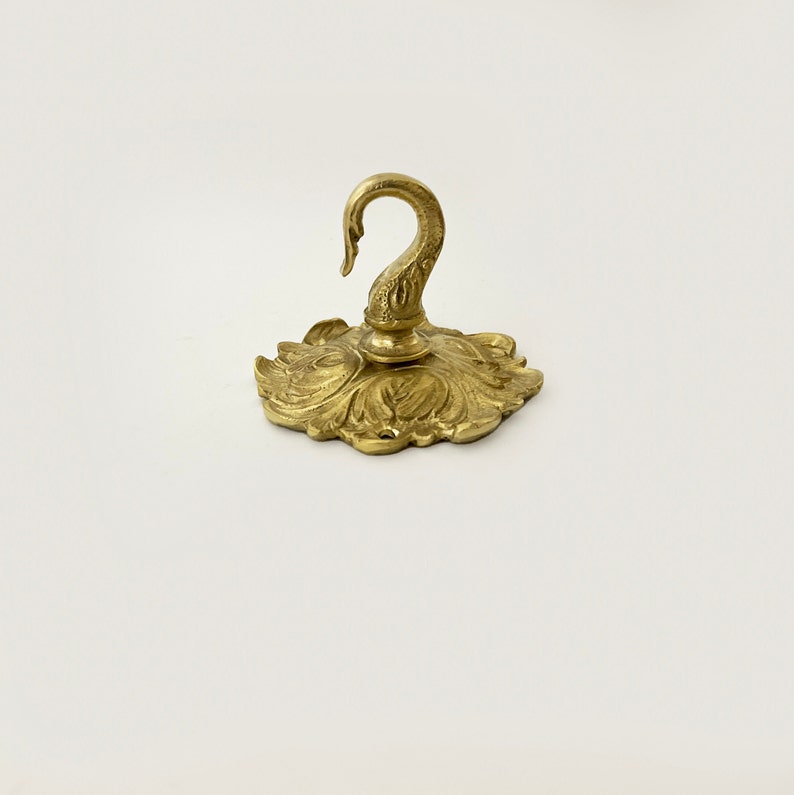 May include: A gold-toned metal hook with a decorative swan design. The hook is attached to a circular base with a floral pattern.