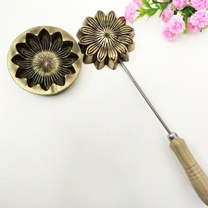 Può includere: Due stampini per biscotti in metallo dorato a forma di fiore. Uno stampino è attaccato a un manico di legno con un'asta di metallo.