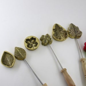 Op de afbeelding: Vijf messingvormen voor het maken van fondantbloemen en -bladeren. De vormen zijn aan houten handvatten bevestigd en hebben verschillende ontwerpen, waaronder een bloem met vijf bloemblaadjes, een blad met een puntige punt en een blad met een afgeronde punt.