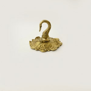 May include: A gold-toned metal hook with a decorative swan design. The hook is attached to a circular base with a floral pattern.