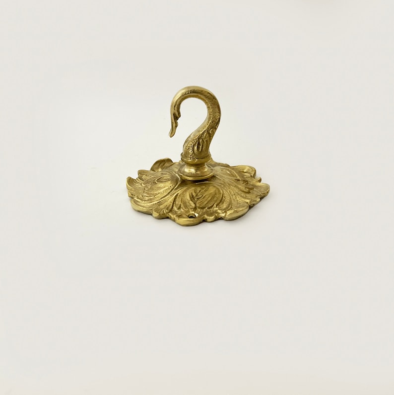 May include: A gold-toned metal hook with a decorative swan design. The hook is attached to a circular base with a floral pattern.