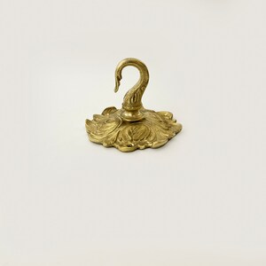 May include: A gold-toned metal hook with a decorative floral design. The hook is attached to a round base with a swan-shaped design.