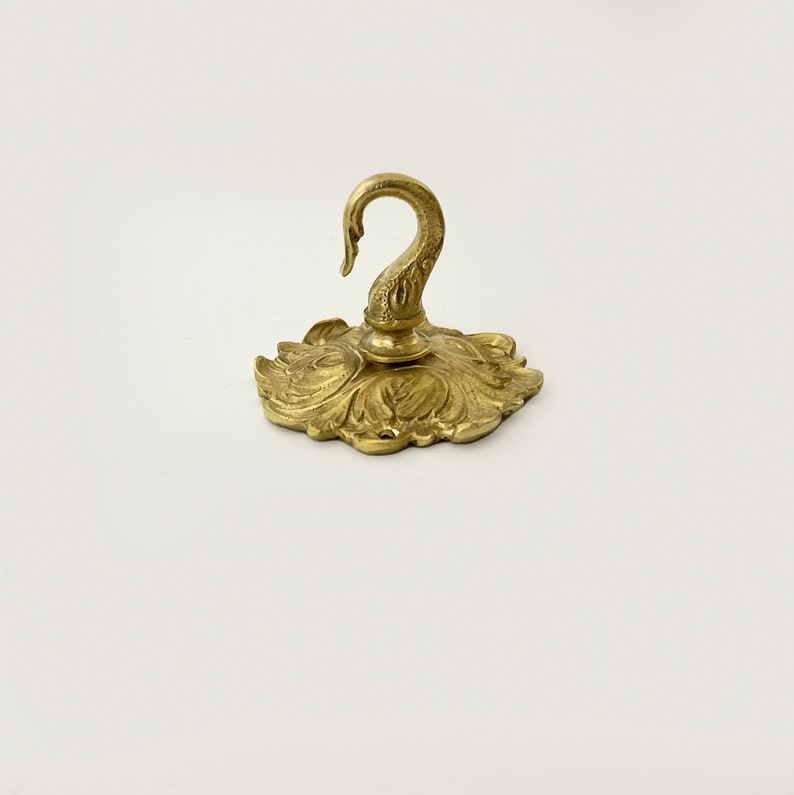 May include: A gold-toned metal hook with a decorative floral design. The hook is attached to a round base with a swan-shaped design.