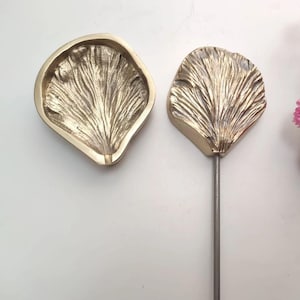 Può includere: Due stampi in metallo color oro per realizzare petali di fiori. Uno stampo è aperto e l'altro è chiuso con un'asta di metallo attaccata.