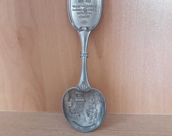 May include: A silver-colored souvenir spoon with a detailed design. The spoon features an ornate handle and a bowl with a scene of people and buildings. The handle has text that reads "Carl Spitzweg 1808-1885".