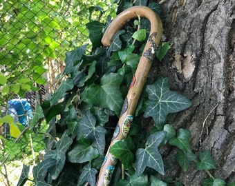 Puede incluir: Un bastón de madera con un mango curvo. El bastón está decorado con pequeñas pegatinas de colores y descansa contra un tronco de árbol cubierto de hiedra verde. El bastón es de color marrón claro.