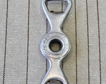 May include: A silver-colored metal bottle opener with a symmetrical, elongated shape featuring two triangular openings at each end and a circular center. The word "VAUGHAN" is embossed in a circular pattern around the central hole of the tool.