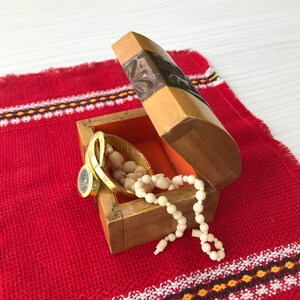 May include: A wooden box with a carved design on the lid is open, revealing a gold bracelet, a watch, and a white beaded necklace. The box is sitting on a red and white patterned fabric.