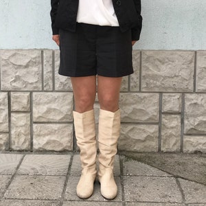 Peut inclure: Une femme portant une veste noire, un chemisier blanc, un short noir et des bottes montantes crème. Elle est debout devant un mur en pierre.