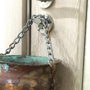 May include: A copper pot with a chain handle, showing signs of age and patina. The pot has a rounded body and a slightly flared rim. The chain is attached to the pot with rivets and hangs from a door handle.