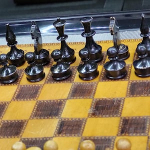 May include: A black and white chess set with black pieces on a wooden chessboard. The board has a light brown and dark brown checkered pattern. The pieces are arranged in a starting position for a game of chess.