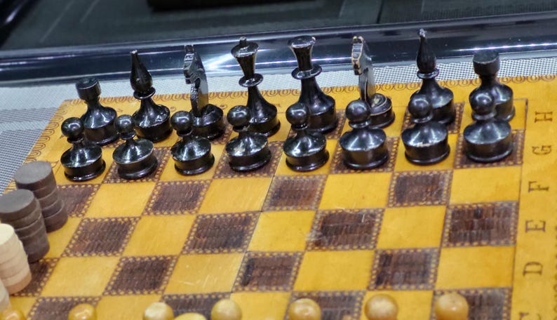 May include: A black and white chess set with black pieces on a wooden chessboard. The board has a light brown and dark brown checkered pattern. The pieces are arranged in a starting position for a game of chess.