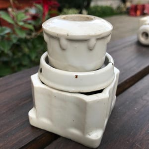 Può includere: Un portafusibile elettrico vintage in ceramica bianca. Il portafusibile è composto da tre sezioni impilate, con una parte superiore cilindrica, una parte centrale circolare e una base quadrata. L'oggetto è posto su una superficie di legno scuro.