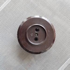 Puede incluir: Una toma de corriente de plástico marrón con tres agujeros redondos. La toma de corriente está sobre una superficie texturizada blanca y gris.