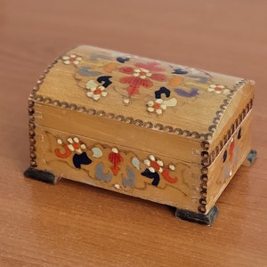 May include: A small wooden box with a hinged lid. The box is decorated with hand-painted floral designs in shades of red, orange, yellow, blue, and green. The box has a scalloped edge and sits on four small feet.