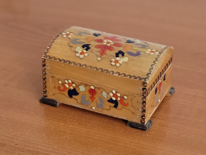 May include: A small wooden box with a hinged lid. The box is decorated with hand-painted floral designs in shades of red, orange, yellow, blue, and green. The box has a scalloped edge and sits on four small feet.