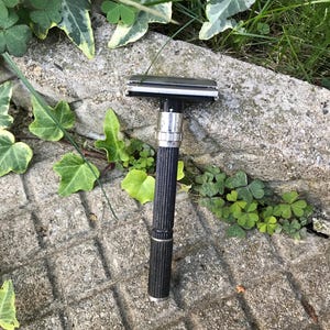 Puede incluir: Una maquinilla de afeitar de seguridad negra y plateada con un mango texturizado. La maquinilla se encuentra sobre un fondo de follaje verde y una superficie de piedra gris. El cabezal de la maquinilla es plateado y el mango es negro con un patrón texturizado.