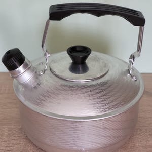 Könnte beinhalten: Ein silberner Aluminium-Wasserkocher mit einem schwarzen Kunststoffgriff und -deckel. Der Wasserkocher hat ein strukturiertes, wellenförmiges Design.