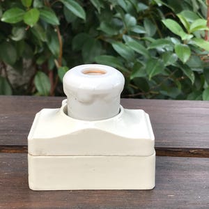 Può includere: Presa elettrica vintage color crema, con parte superiore arrotondata e base rettangolare. La parte superiore ha un'apertura circolare. La presa è in ceramica o porcellana. Lo sfondo presenta un fogliame verde.