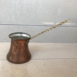 Op de afbeelding: Een koperen koffiepot met een gehamerde afwerking en een messing handvat. Het handvat heeft een decoratief ontwerp.