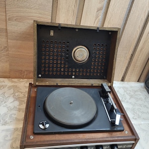 May include: A vintage wooden record player with a black turntable and a speaker in the lid. The record player has a silver trim and the word "RIGA" is printed on the front.