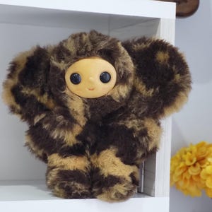 Op de afbeelding: Een pluche speelgoed met een rond, lichtgekleurd gezicht en grote zwarte ogen. Het lichaam is bedekt met bruin en tan patroonbont, met grote, afgeronde oren. Het speelgoed staat op een witte plank.