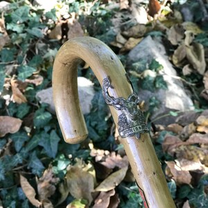 Puede incluir: Un bast&oacute;n de madera de color claro con un mango curvo. El bast&oacute;n presenta una banda decorativa de metal plateado con un emblema de ciervo y la palabra "KONIGSBURG". El bast&oacute;n se encuentra sobre un fondo de follaje verde y marr&oacute;n.